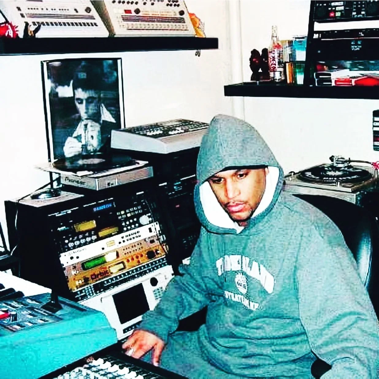 DJ Clear in his studio in the hardware era: drum machines lined up on the top shelf, a rack of outboard gear and samplers at his desk, a turntable within arm's reach, a framed black-and-white portrait on the wall
