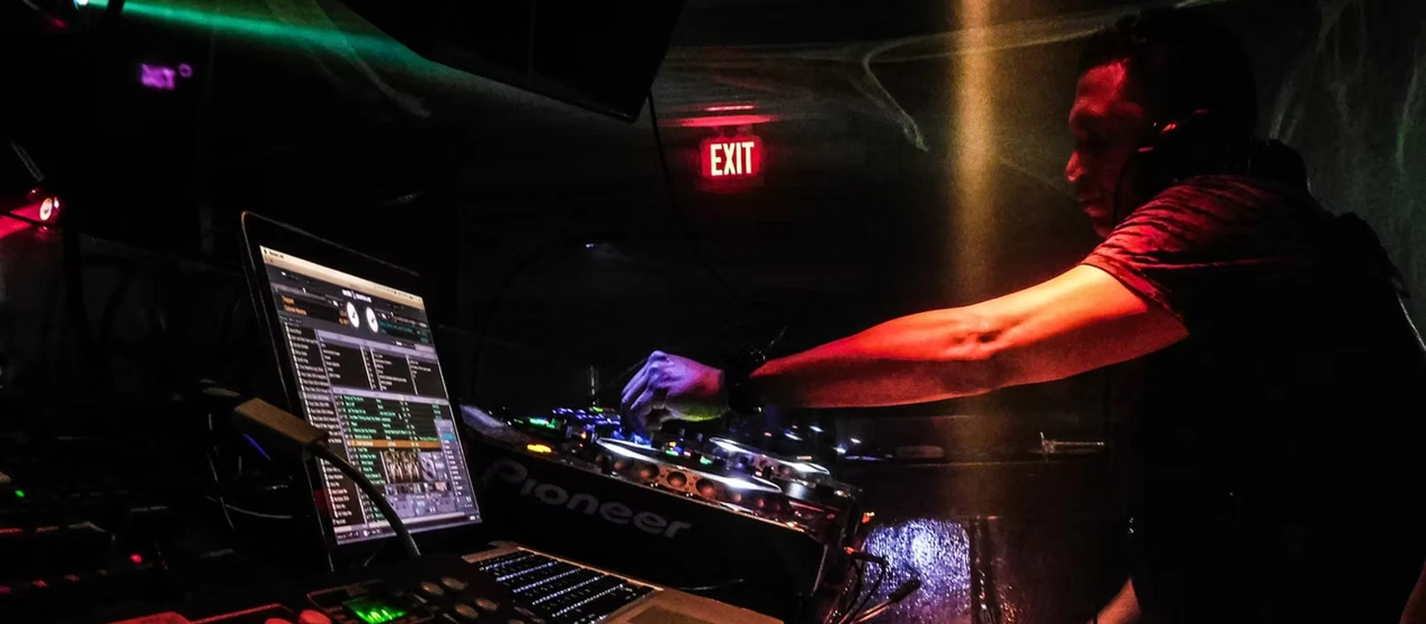 DJ Clear mid-set in a club under red light, headphones on, reaching across the Pioneer decks beside a laptop running Serato
