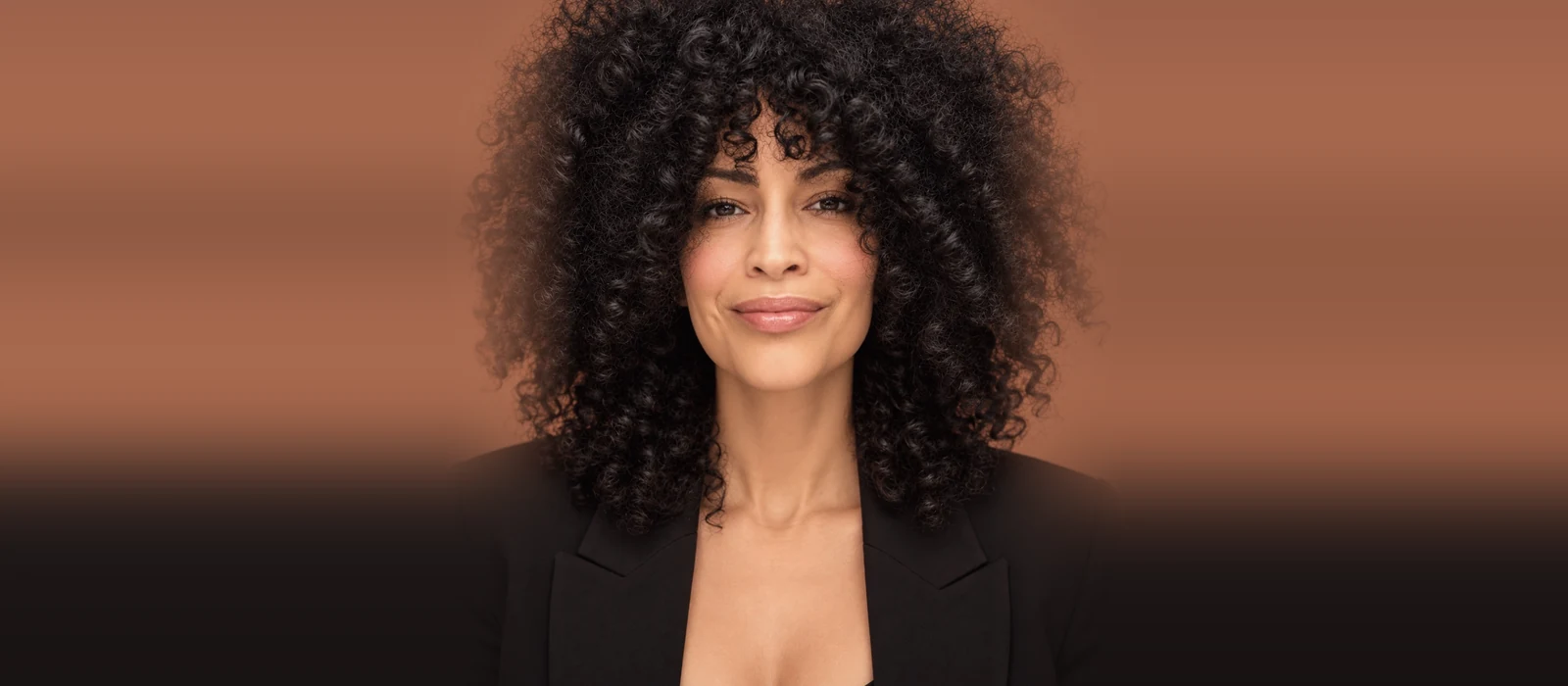 Portrait of DJ Tabu smiling at the camera, with voluminous curly black hair and a black blazer against a terracotta backdrop