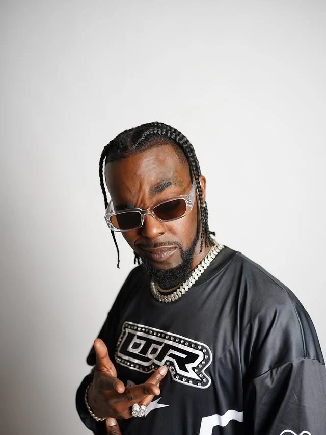 Roscoe Dash in clear-framed sunglasses, braids and a silver chain, pointing toward the camera