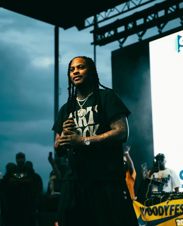 Waka Flocka Flame on stage with long braids, a black graphic tee and a chain, holding a microphone